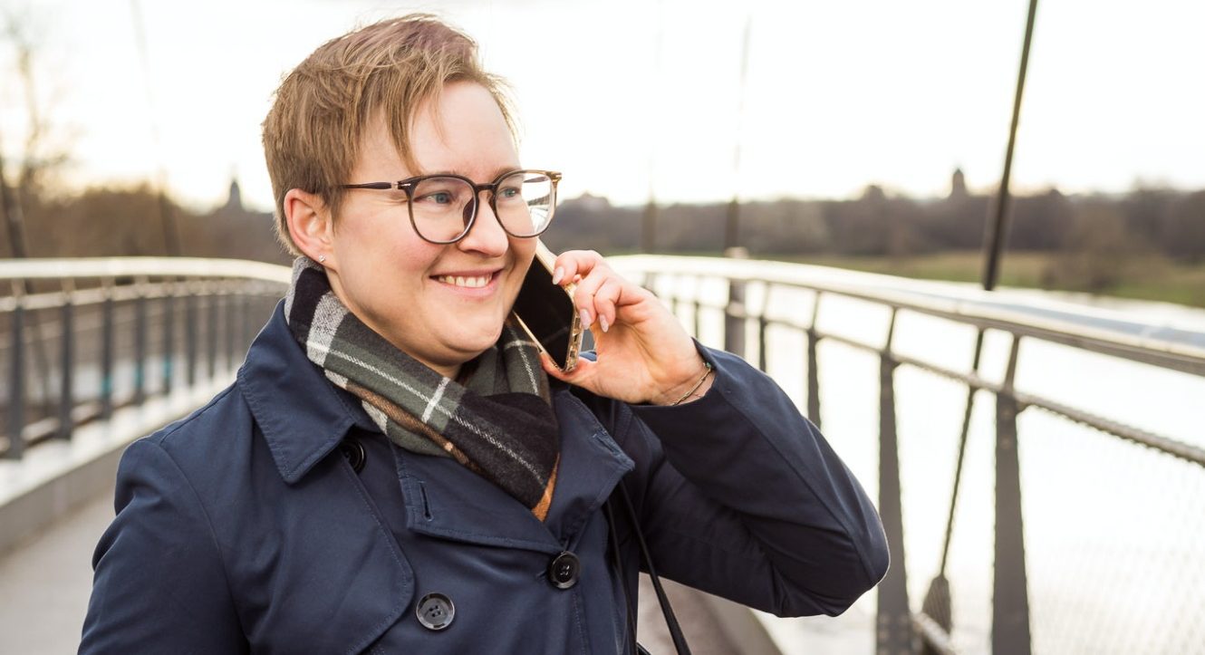 Junge Person mit Brille und Schal, die lächelt und telefoniert auf einer Brücke.
